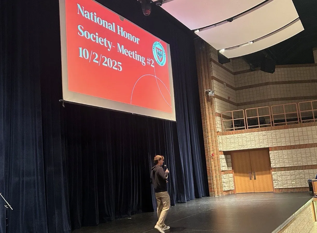 Maclane Zich speaking into a microphone on the Benet Academy stage beneath a National Honor Society meeting slide